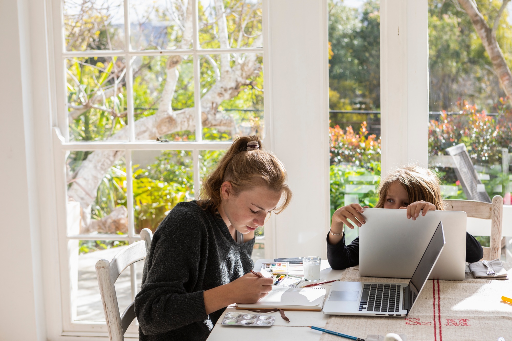 Teenage girl painting with watercolours at a table, and a boy on a laptop