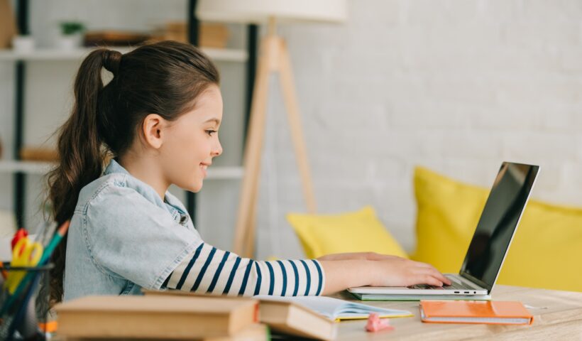 side view of adorable child using laptop while sitting at desk with books and doing homework