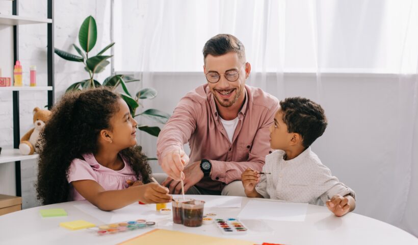 smiling caucasian teacher in eyeglasses and african american kids drawing pictures with paints
