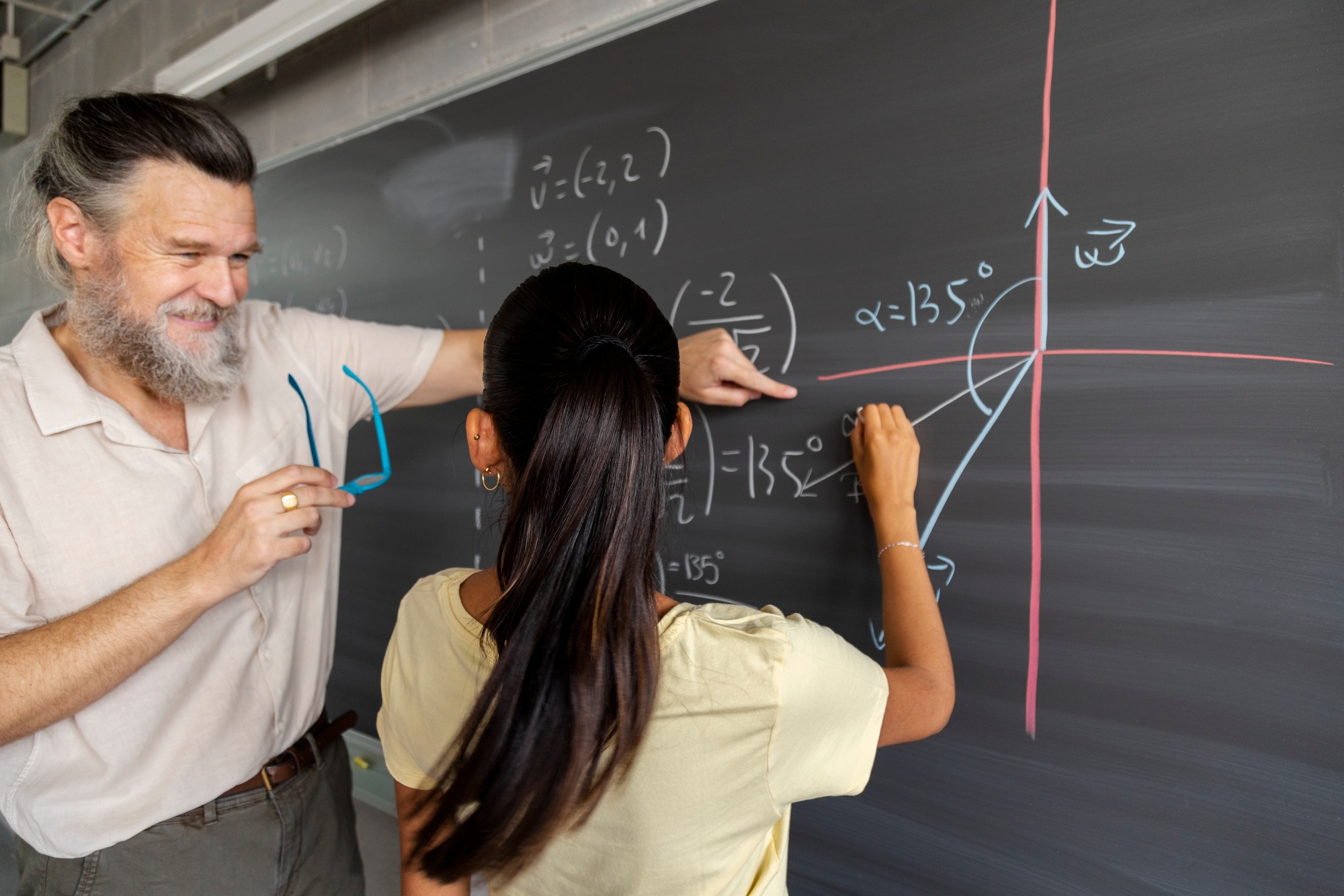 Smiling male teacher shows female teen high school student math problem resolution on blackboard.