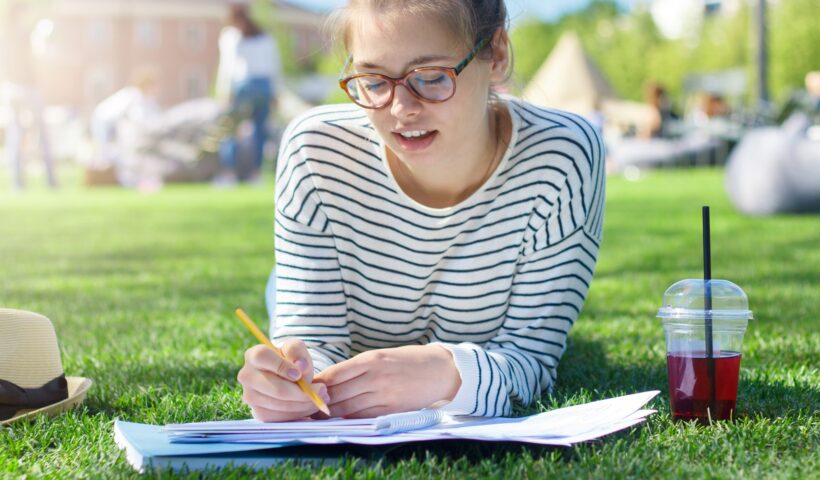 Student girl lying on green grass with notebook open in front, texting messages