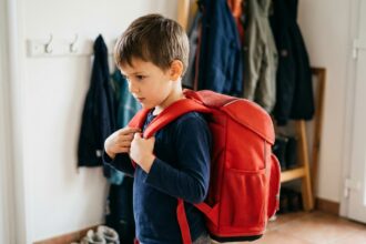 Jeune garçon essayant un cartable ergonomique rouge dans l'entrée de sa maison avant la rentrée scolaire